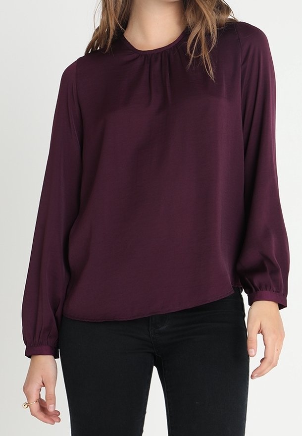 Femme portant un chemisier à manches longues, de couleur violet foncé, avec un col et des poignets froncés, associé à un pantalon noir ajusté.