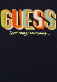 Granatowy materiał z napisem "GUESS" w kolorowej, wielowarstwowej haftowanej aplikacji oraz hasłem "Wielkie rzeczy nadchodzą..." poniżej.