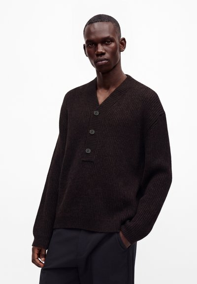 Homme portant un pull en maille côtelée marron foncé avec trois boutons sur le col en V et un pantalon sombre, debout devant un fond blanc.