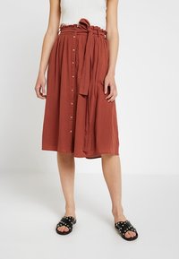 Woman wearing a rust-colored, button-front midi skirt with a tied waist belt, a white sleeveless top, and black studded slide sandals.