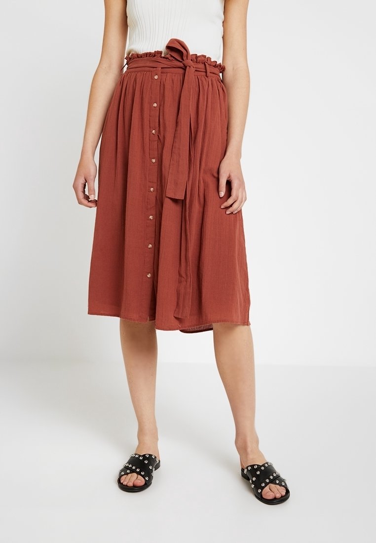 Woman wearing a rust-colored, button-front midi skirt with a tied waist belt, a white sleeveless top, and black studded slide sandals.