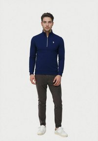 Navy blue zip-up sweater with long sleeves and ribbed cuffs, paired with dark gray pants and white sneakers. Subtle embroidered logo on chest.