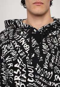 Sweat à capuche noir avec un impression en écriture blanche audacieuse et divers mots. Conçu avec une capuche à cordon et un tissu texturé.