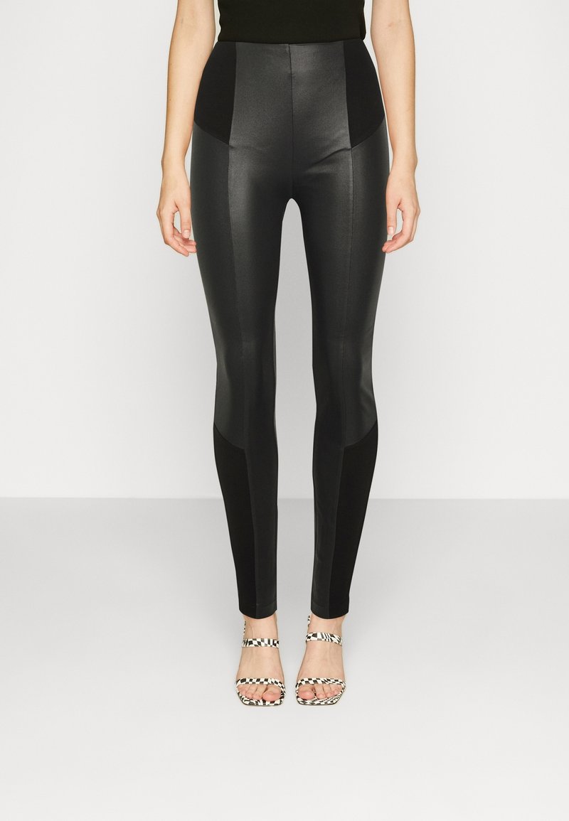 Femme portant des leggings taille haute en similicuir noir et des talons ouverts à motif zèbre, debout devant un fond clair uni.