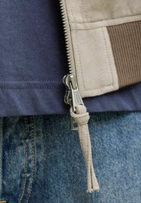 Gros plan sur la fermeture éclair d'une veste beige avec un tire-zip en tissu, portée sur une chemise marine et un jean bleu.