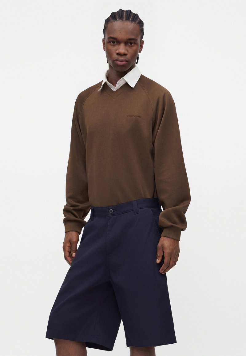 Mand med flettet hår iført en brun langærmet sweater over en hvid skjorte med krave og navyblå knælange shorts.