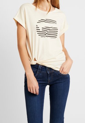 Femme portant un t-shirt écru aux manches retroussées avec un logo cercle à rayures noires et un nœud noué, associé à un jean bleu foncé.