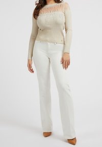 Béžový žebrovaný svetr s průhledným výstřihem a flitrovými detaily, kombinovaný s bílými rozšířenými džíny a světle hnědými lodičkami s špičatou špičkou.