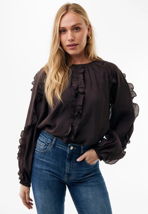 Blonde Frau trägt eine schwarze langärmelige Rüschenbluse und blaue hoch taillierte Jeans mit einer Hand in der Tasche vor weißem Hintergrund.