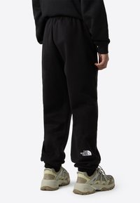 Svarte joggebukser med elastisk midjebånd og ribbede ankler, med en hvit logo på venstre ben. Brukes med beige og grå joggesko.