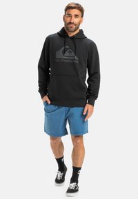 Fekete kapucnis pulóver Quiksilver logóval, kenguruzsebből áll, kék sorttal, fekete zoknikkal és fekete cipővel párosítva. Kényelmes, laza viselet.