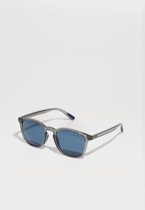 Óculos de sol cinzentos translúcidos com lentes azuis, uma forma angular e acabamentos em prata nas hastes. Marcação notável na lente.