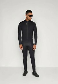 Mann iført svart sykkeljakke, svarte sykkelbukser, svarte sykkelsko og mørke solbriller, står mot en enkel hvit bakgrunn.