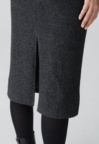 Jupe côtelée gris foncé avec une fente centrale au dos, portée avec des collants noirs et des bottines noires, une main reposant sur la hanche.