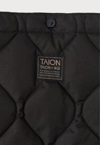 Tecido preto acolchoado com uma textura brilhante, apresentando uma etiqueta de marca proeminente em letras douradas e um botão preto na parte superior.