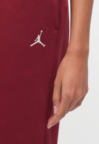 Κοντινό πλάνο ενός χεριού που ακουμπά δίπλα σε βυσσινί υφασμάτινο παντελόνι με μικρό λευκό λογότυπο Jumpman κεντημένο κοντά στη ραφή της τσέπης.