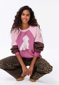 Růžová a hnědá mikina s velkým bílým grafickým motivem zvířete a textem, doplněná hnědými kalhotami s leopardím potiskem. Měkká tkanina a uvolněný střih.