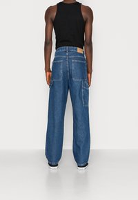 Farkku cargo-housut, tummansininen, leveä lahje, jossa on useita taskuja ja vaaleanruskea etiketti takana. Yhdistetty mustan hihattoman paidan ja tennareiden kanssa.