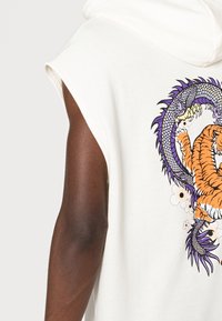 Ärmlös vit hoodie med en färgglad grafik av en tiger som är sammanflätad med en drake, accentuerad med blommor och djärva konturer.