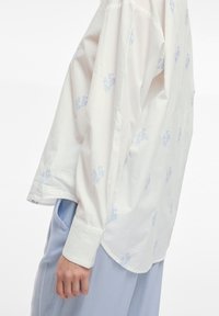 Camicia bianca a maniche lunghe in cotone leggero con ricami floreali azzurri. Orlo arrotondato e polsini con bottone, abbinata a pantaloni azzurri.