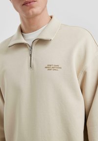 Felpa beige con zip e colletto alto, con testo ricamato in marrone: "NON MI IMPORTA DI NULLA, SOLO RILASSATI." Texture morbida.