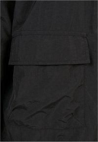 Gros plan d'une poche en tissu noir avec un rabat rectangulaire et des coutures visibles sur un vêtement.