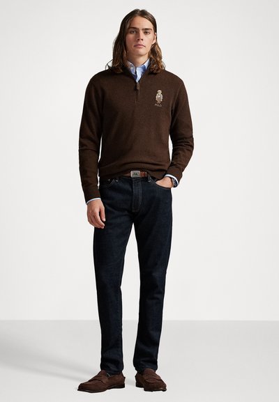 Rjav zip-up pulover z izvezenim logotipom, v kombinaciji s temnimi kavbojkami in rjavimi loaferji. Model stoji z dolgimi lasmi in rokami v žepih.