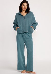 Teal lounge sæt bestående af en lynlåsjakke uden krave med sidelommer og elastiske manchetter, parret med løstsiddende bukser med snoretræk.