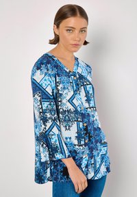 Bluse in Blau und Weiß mit Muster, V-Ausschnitt, Dreiviertelärmeln und Knopfverschluss vorne. Sie weist florale und geometrische Designs auf.