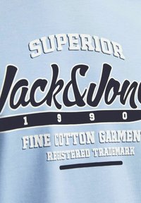 Vaaleansininen puuvillapaita, jossa on rohkeaa musta-valkoista tekstiä: "SUPERIOR Jack&Jones 1990 HENKILÖKOHTAISET PUUVILLATUNNISTEET, REKISTERÖITY KAUPPAKUNTO."