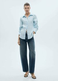 Camicia di lino azzurro chiaro con bottoni frontalmente e maniche lunghe, abbinata a jeans scuri a gamba larga. Il look è completato da sandali neri con cinturini.