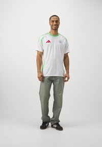 Jersey bianco a maniche corte con dettagli verdi, caratterizzato da un emblema e logo Adidas rosso sul petto, abbinato a jeans blu e sneaker nere.