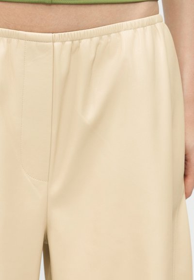 Κοντινό πλάνο μπεζ παντελονιού από συνθετικό δέρμα με ελαστική μέση, φορεμένο από ένα άτομο, που δείχνει λεπτομέρειες ραφής και ομαλή υφή.