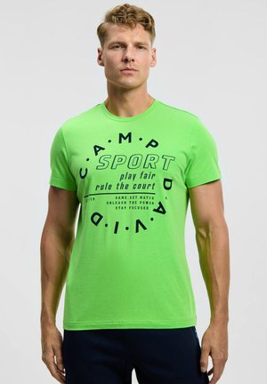 Young man wearing bright green t-shirt with black sports slogan text standing against plain light background.