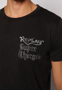 Czarna bawełniana koszulka z okrągłym dekoltem, z srebrnym napisem: "Replay Super Charged" w różnych stylizowanych czcionkach i rozmiarach.