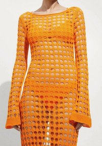 Femme portant une robe en crochet orange à manches longues avec de grands trous circulaires et un sous-vêtement assorti visible en dessous.