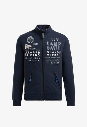 Navy zip-up jacket with a high collar, featuring white text and nautical graphics. Includes two front pockets with blue zipper pulls.