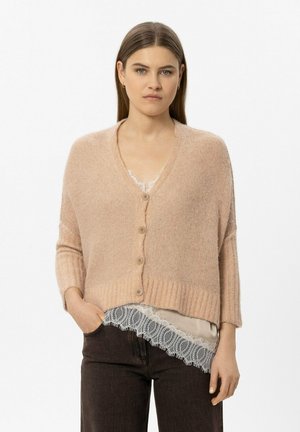 Woman with straight brown hair wearing a beige buttoned cardigan over a lace-trimmed top and dark pants, standing with one hand in pocket.