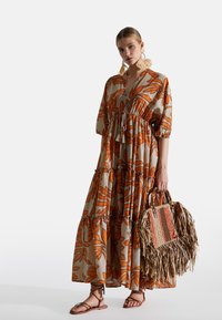 Maxikleid in Beige mit orangefarbenem Blätterdruck, V-Ausschnitt, gestuftem Design und taillierter Taille. Kombiniert mit einer geflochtenen Tasche und flachen Sandalen.