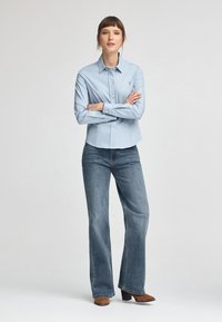 Camicia button-up blu chiaro con colletto a punta, abbinata a jeans a gamba larga blu sbiaditi e stivali con tacco alla caviglia marroni. Dettagli di cucitura notevoli.