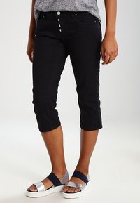 Henkilö, joka on pukeutunut mustiin lyhyisiin housuihin, joissa on napit, sekä hopeisiin ja tummansinisiin korkokenkiin, seisoo valkoisella lattialla.