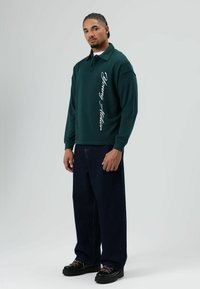 Donkergroene polo met lange mouwen en zijkanttekst, gecombineerd met losse donkere denimbroek en zwarte schoenen met roze veters en gestructureerde zolen.