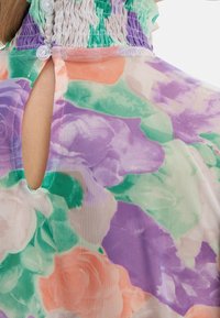 Blouse florale transparente présentant une palette de couleurs pastel avec des roses violettes, vertes et pêches, un col smocké et un détail en forme de trou de serrure à l'arrière.