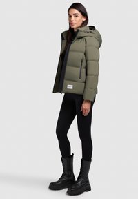 Khaki pufferjakke med hætte, udstyret med en sort lynlås og sidelommer. Kombineret med sorte leggings og knæhøje snørestøvler.