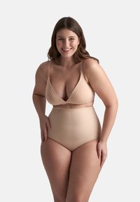 Donna sorridente con lunghi capelli castani, che indossa un'intimo beige a vita alta abbinato a un reggiseno, in piedi su uno sfondo semplice.