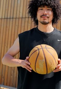 Black sleeveless athletic shirt; a man holds a brown basketball showing logo and seams; wooden fence in the background.
