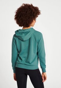 Teal hoodie med dragsko, ribbade ärmslut och nederkant samt mjuk textur. Har en avslappnad passform och raglanärmar. Svarta leggings synliga.