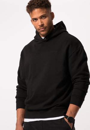 Zwarte hoodie met een gestructureerde afwerking, voorzien van een voorzak, geribbelde manchetten en een subtiele logo. Draagt over een wit shirt en gecombineerd met donkere broek.