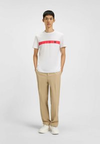 T-shirt bianca con una striscia rossa orizzontale e logo "HUGO", abbinata a pantaloni beige e sneakers bianche. Design semplice e collo rotondo.