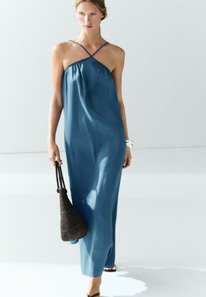 Frau trägt ein blaues ärmelloses Maxikleid und hält eine schwarze geflochtene Tasche, sie geht in einem minimalistisch hellen Raum mit weißen Wänden und Boden.
